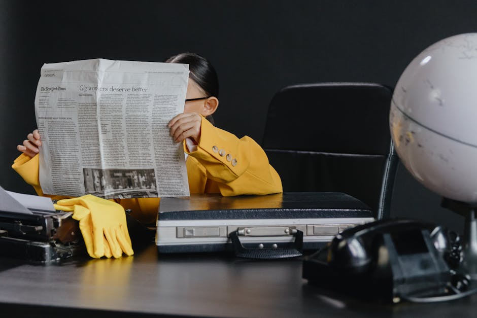 A young girl in a yellow suit reading a newspaper at a desk with office objects.